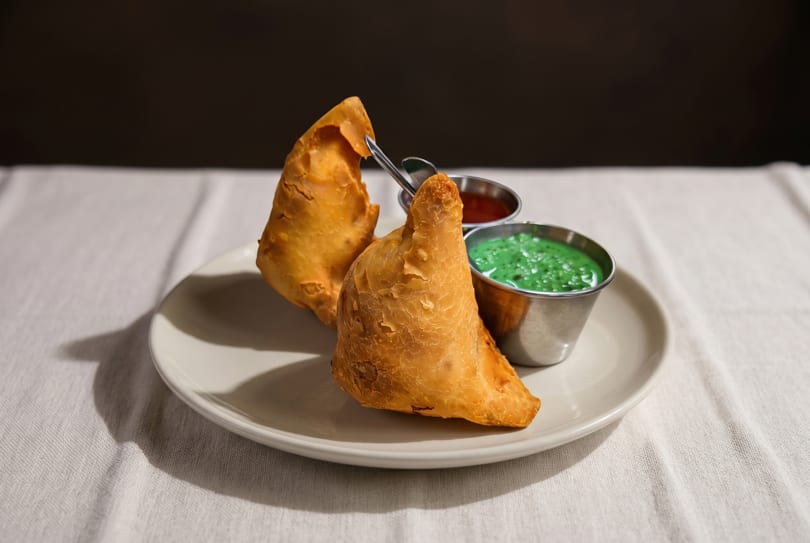 Two golden-brown samosas on a white plate with green and red dipping sauces.