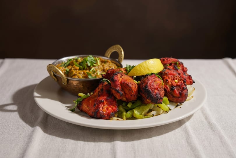 Tandoori chicken with grilled vegetables and a side of curry, garnished with lemon and cilantro.